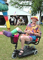Photo: A smiling man in colorful clothing. He is sitting in a scooter with his leg in a decorated cast and is clearly enjoying his visit to this festival.